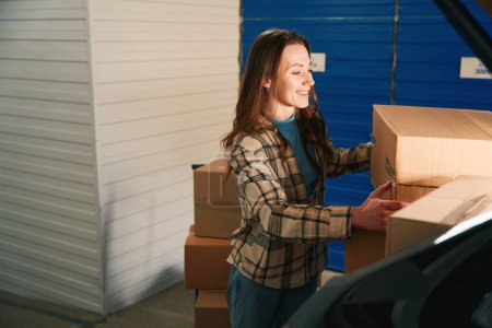 Téléchargez les photos : Happy Lady met des boîtes en carton dans le coffre d'une voiture dans un entrepôt avec unité de self-stockage - en image libre de droit