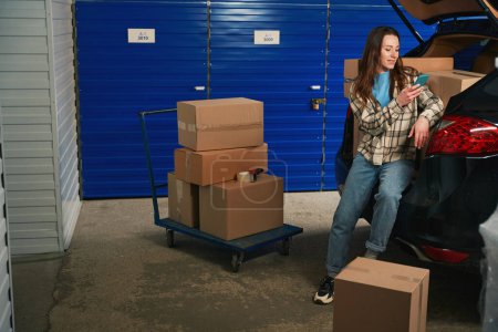 Photo for Young woman is calling with mobile and standing near the trunk of car in a storage warehouse. Cardboard boxes with things are on platform truck and in the trunk of car - Royalty Free Image
