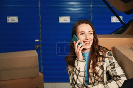 Photo for Smiling lady is calling with mobile and sitting in the trunk of car in a storage warehouse. Cardboard boxes with things are in the trunk of car - Royalty Free Image