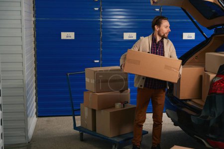 Téléchargez les photos : Le gars met de grandes boîtes en carton du camion à plate-forme dans le coffre d'une voiture dans un entrepôt de stockage - en image libre de droit