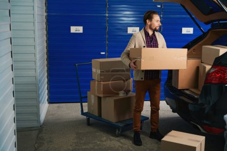Foto de Guy está poniendo grandes cajas de cartón de camión plataforma en el maletero de un coche en el almacén de almacenamiento - Imagen libre de derechos