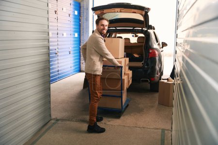 Téléchargez les photos : Un homme souriant roule le camion à plate-forme avec de grandes boîtes en carton vers la voiture dans un entrepôt de stockage - en image libre de droit
