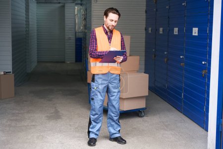 Photo for Worker guy is standing and writing notes with tablets for writing into storage warehouse - Royalty Free Image