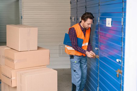 Photo for Guy is opening the storage room in the warehouse and holding tablets for writing. Many big cardboard boxes are standing near - Royalty Free Image
