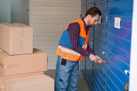 Photo for Man is opening the storage room in the warehouse and holding tablets for writing. Many big cardboard boxes are standing near - Royalty Free Image