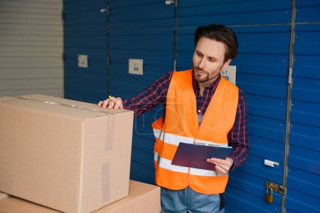 Téléchargez les photos : Jeune homme est debout près de grandes boîtes en carton et de vérifier l'emballage dans l'entrepôt de stockage - en image libre de droit
