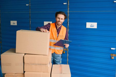 Photo for Smiling guy is standing near big cardboard boxes and checking the packaging in storage warehouse - Royalty Free Image