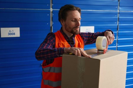 Téléchargez les photos : Jeune homme travailleur emballe grande boîte en carton avec pistolet à ruban et scotch dans l'entrepôt - en image libre de droit