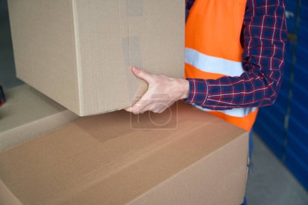 Foto de Chico en ropa de trabajo está sosteniendo una gran caja de cartón en las manos en el almacén con unidad de almacenamiento - Imagen libre de derechos