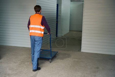 Téléchargez les photos : L'homme en salopette pour les travailleurs roule le camion à plate-forme vide dans un entrepôt de stockage - en image libre de droit