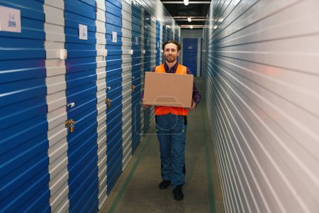 Photo for Happy guy is holding a big cardboard box and standing in overalls for the workers in storage warehouse - Royalty Free Image