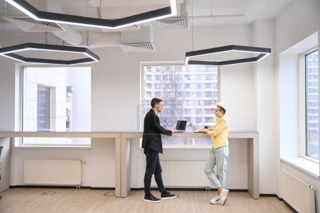 Photo for Workteam discussing start-up project standing near counter with laptops in open space hub with big windows - Royalty Free Image