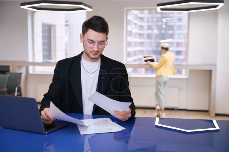 Téléchargez les photos : Jeune homme avec ordinateur portable regardant à travers les documents, travaillant dans un espace de coworking - en image libre de droit