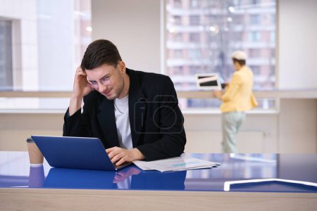 Foto de Pensativo joven escribiendo en el ordenador portátil en el espacio de coworking, pensando en la estrategia de negocios, plazo - Imagen libre de derechos