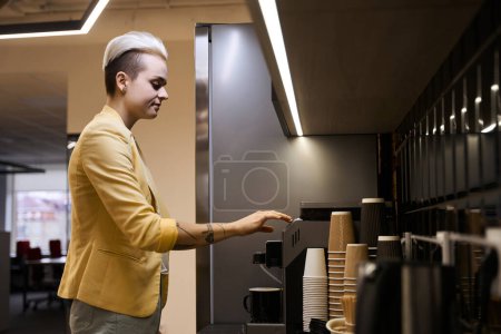 Foto de Mujer analista pulsando el botón para preparar café en la máquina de café en la cocina de la oficina, hora del almuerzo - Imagen libre de derechos