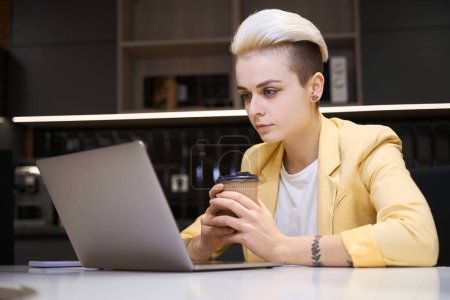 Téléchargez les photos : Programmeuse concentrée buvant du café et travaillant sur un ordinateur portable dans un club ouvert, vérifiant le code Web - en image libre de droit