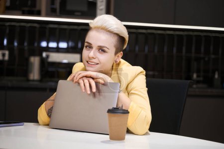 Téléchargez les photos : Femme attrayante appuyée sur un ordinateur portable, posant caméra assise sur le lieu de travail dans un espace de coworking, développant son site Web - en image libre de droit