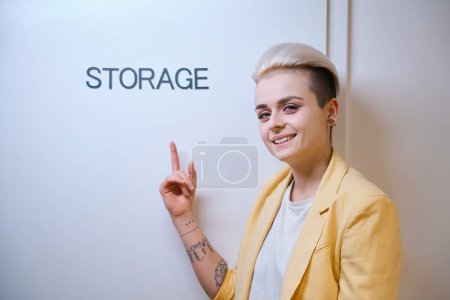Photo for Smiling woman pointing finger at storage sign on door, room for office stuff safekeeping, authorized personnel only - Royalty Free Image