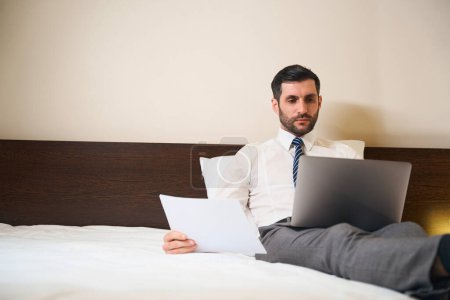 Foto de Hombre guapo medio sentado en almohadas, situado en la cama con documentos de trabajo y un ordenador portátil - Imagen libre de derechos