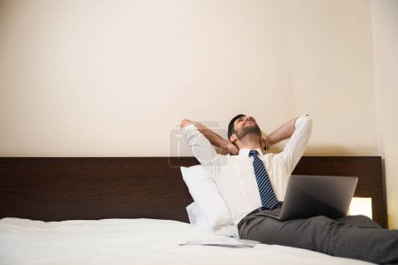 Foto de Hombre, medio sentado en almohadas, sentado en la cama con los brazos sobre la cabeza, se detuvo en el trabajo en el portátil - Imagen libre de derechos