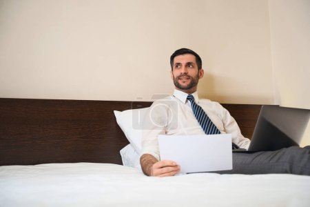Téléchargez les photos : Homme à moitié assis sur des oreillers, assis sur un lit avec des documents de travail et un ordinateur portable - en image libre de droit