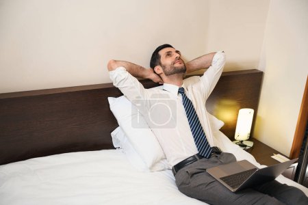 Photo for Man lies on the bed with his hands thrown over his head, he paused in work on the laptop - Royalty Free Image