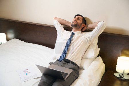 Téléchargez les photos : Homme fermé les yeux de fatigue, il, sans se déshabiller, travaille sur un ordinateur portable dans un lit confortable - en image libre de droit