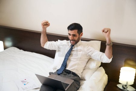 Foto de Hombre se regocija en sus logros, él, sin desnudarse, trabaja en un ordenador portátil en una cama cómoda, - Imagen libre de derechos