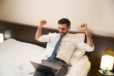 Foto de Hombre, sin desnudarse, trabaja en un ordenador portátil en una cama cómoda, se regocija en sus logros - Imagen libre de derechos