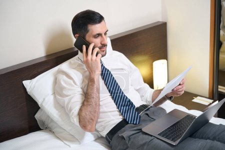 Foto de Hombre con ropa de oficina está estudiando documentos de trabajo y hablando por teléfono, se encuentra en una cama grande - Imagen libre de derechos