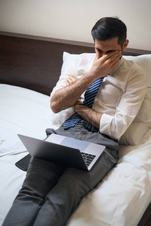 Foto de El hombre cansado está luchando con el sueño mientras trabaja en documentos, que se encuentra en una cama grande - Imagen libre de derechos