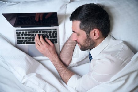 Téléchargez les photos : Homme fatigué s'est endormi, sans se déshabiller, au travail sur l'ordinateur portable, il s'est installé sur un lit confortable - en image libre de droit