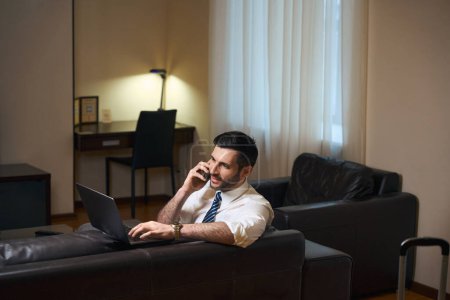 Téléchargez les photos : Homme en tenue de travail travaille dans l'espace détente de la chambre d'hôtel, il utilise ordinateur portable et téléphone - en image libre de droit