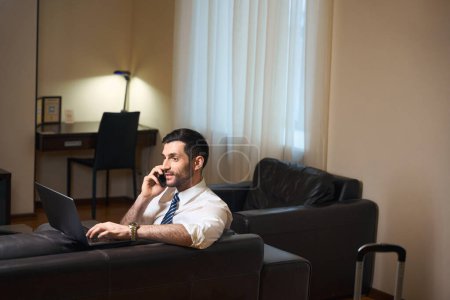 Téléchargez les photos : Homme en tenue de travail est situé sur un canapé en cuir avec ordinateur portable et un téléphone, il travaille dans la zone de détente - en image libre de droit