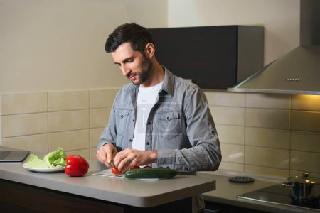 Photo for Middle-aged man cuts vegetables for salad, male is located in the kitchen area of the hotel room - Royalty Free Image