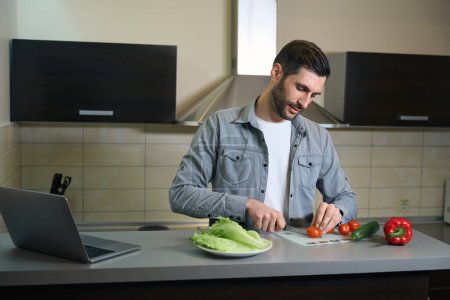 Foto de Hombre en ropa casual corta verduras para salat, el hombre se encuentra en la zona de cocina de la habitación de hotel - Imagen libre de derechos