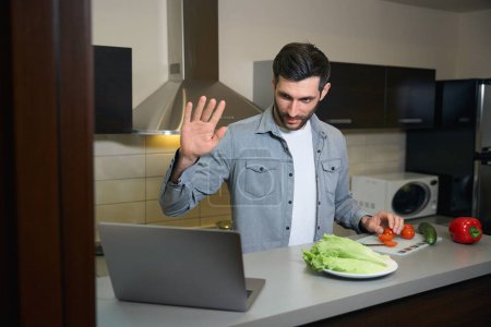 Foto de Hombre en ropa casual prepara ensalada y se comunica por ordenador portátil, el hombre se encuentra en la zona de cocina de la habitación del hotel - Imagen libre de derechos