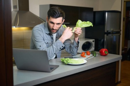 Téléchargez les photos : Homme prépare une salade de légumes et communique par ordinateur portable, l'homme est situé dans la cuisine de la chambre d'hôtel - en image libre de droit