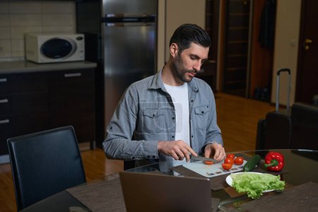 Téléchargez les photos : Homme coupe une tomate pour une salade de légumes, à côté de lui sur la table est un ordinateur portable - en image libre de droit