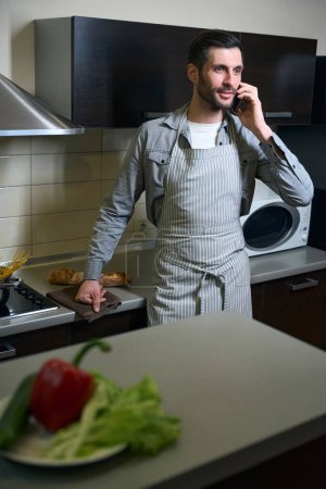 Photo for Brunette in a chefs apron stands in the kitchen with a phone, a plate of vegetables is on the table - Royalty Free Image