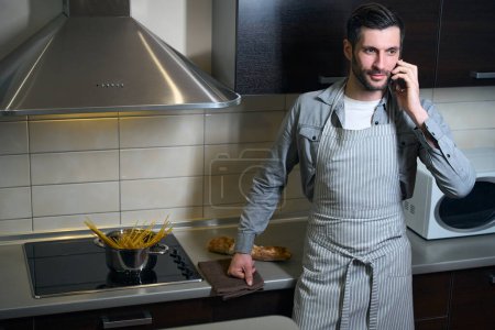 Téléchargez les photos : Homme dans un tablier de chefs est debout dans la cuisine avec un téléphone, pot de spaghettis est sur la cuisinière - en image libre de droit