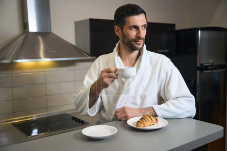Photo for Hotel guest stands with a cup of coffee in the kitchen area, in front of him is saucer with croissant - Royalty Free Image