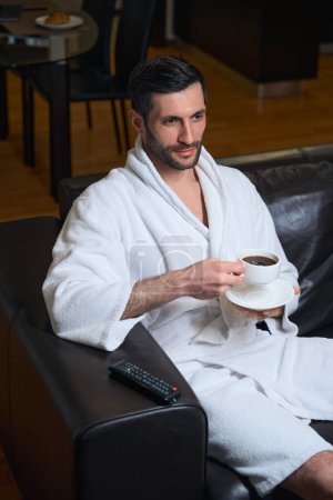 Photo for Handsome man enjoys a cup of morning coffee, the male is located in recreation area, next to the TV remote - Royalty Free Image