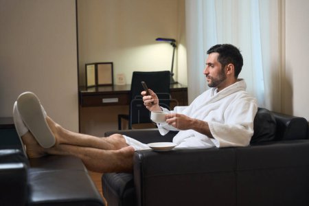 Foto de El huésped del hotel en un albornoz se sienta en una zona de relajación con una taza de café, se comunica con un teléfono móvil - Imagen libre de derechos