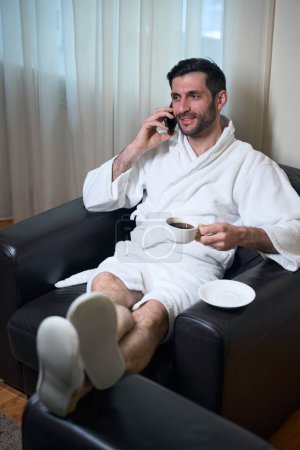 Téléchargez les photos : Homme non rasé en peignoir est assis dans un fauteuil en cuir confortable avec une tasse de café, il parle sur un téléphone mobile - en image libre de droit