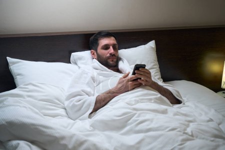 Téléchargez les photos : L'homme fatigué se repose au lit sur des oreillers souples, il a un téléphone portable dans ses mains - en image libre de droit