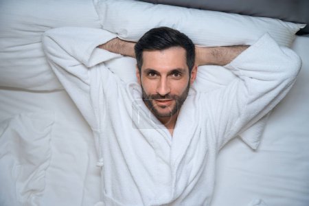 Téléchargez les photos : Satisfait homme reposé se trouve au lit dans une chambre d'hôtel, il est dans un peignoir confortable - en image libre de droit