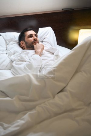 Foto de El hombre está descansando en una cama sobre almohadas suaves en una habitación de hotel, él está en un albornoz - Imagen libre de derechos