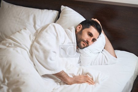 Téléchargez les photos : Fatigué invité de l'hôtel se trouve au lit sur des oreillers doux, il est dans un peignoir - en image libre de droit