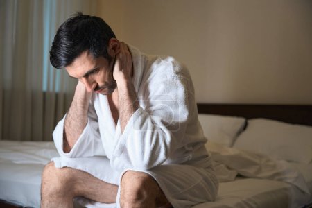 Téléchargez les photos : Homme souffrant assis sur un grand lit dans une chambre d'hôtel, il souffre - en image libre de droit